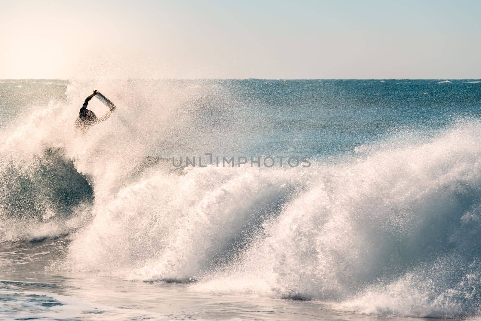 man surfing jumps a great wave that breaks with a lot of power and energy, the sunlight reflects golden on the turquoise surface of the sea and in the plumes of water that the wind raises
