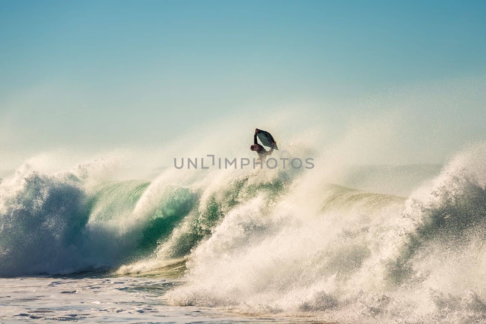 surfer jumping a big wave that breaks with a lot of energy and power, sunlight reflects golden on the turquoise surface of the sea and in the huge pile of foam, the wind rises plumes of water