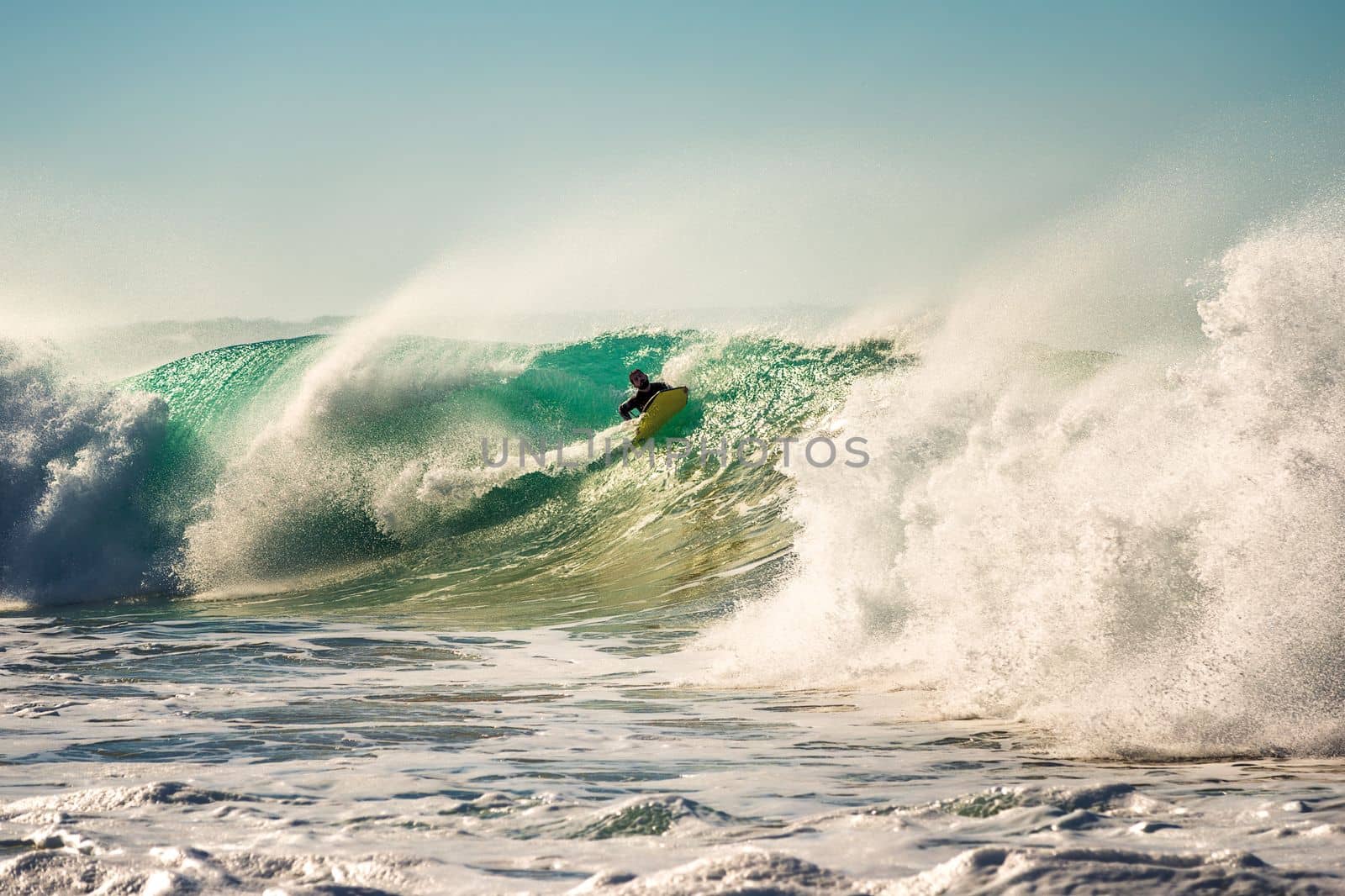 man surfing a big wave that breaks with a lot of energy and power, sunlight reflects golden on the turquoise surface of the sea and in the huge pile of foam, the wind rises plumes of water