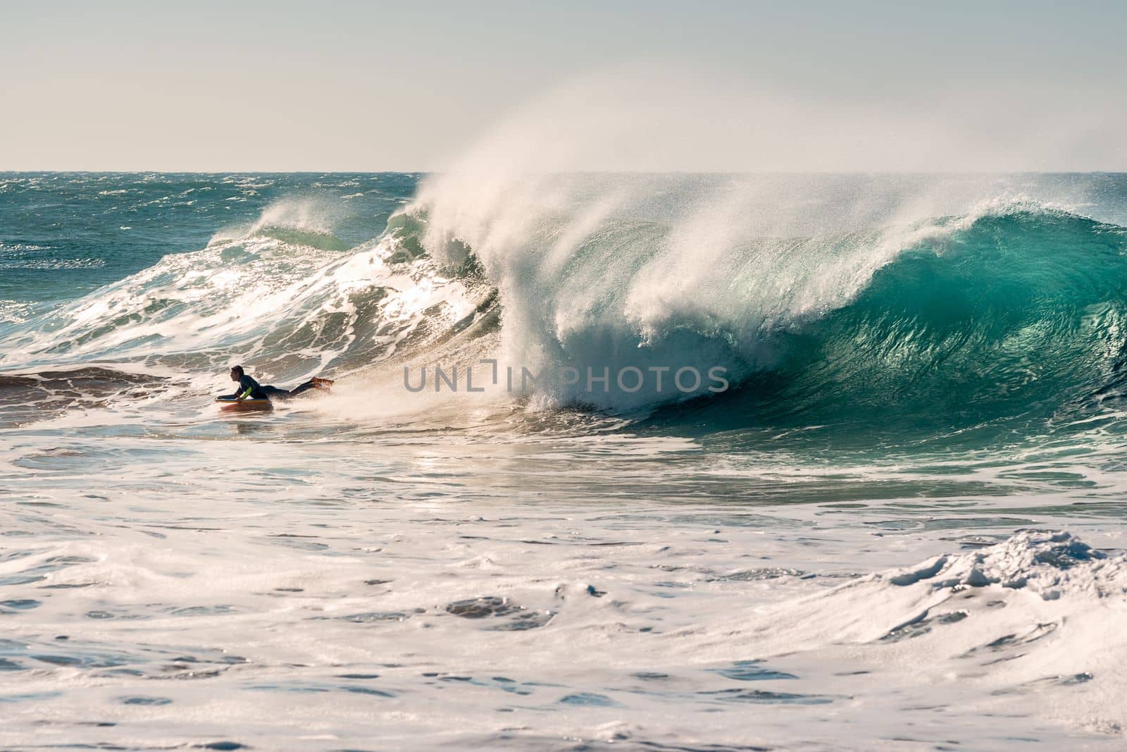 man surfing a huge wave that chases him, the sunlight reflects golden on the turquoise surface of the sea and on the foam and the plumes of water that the wind blows