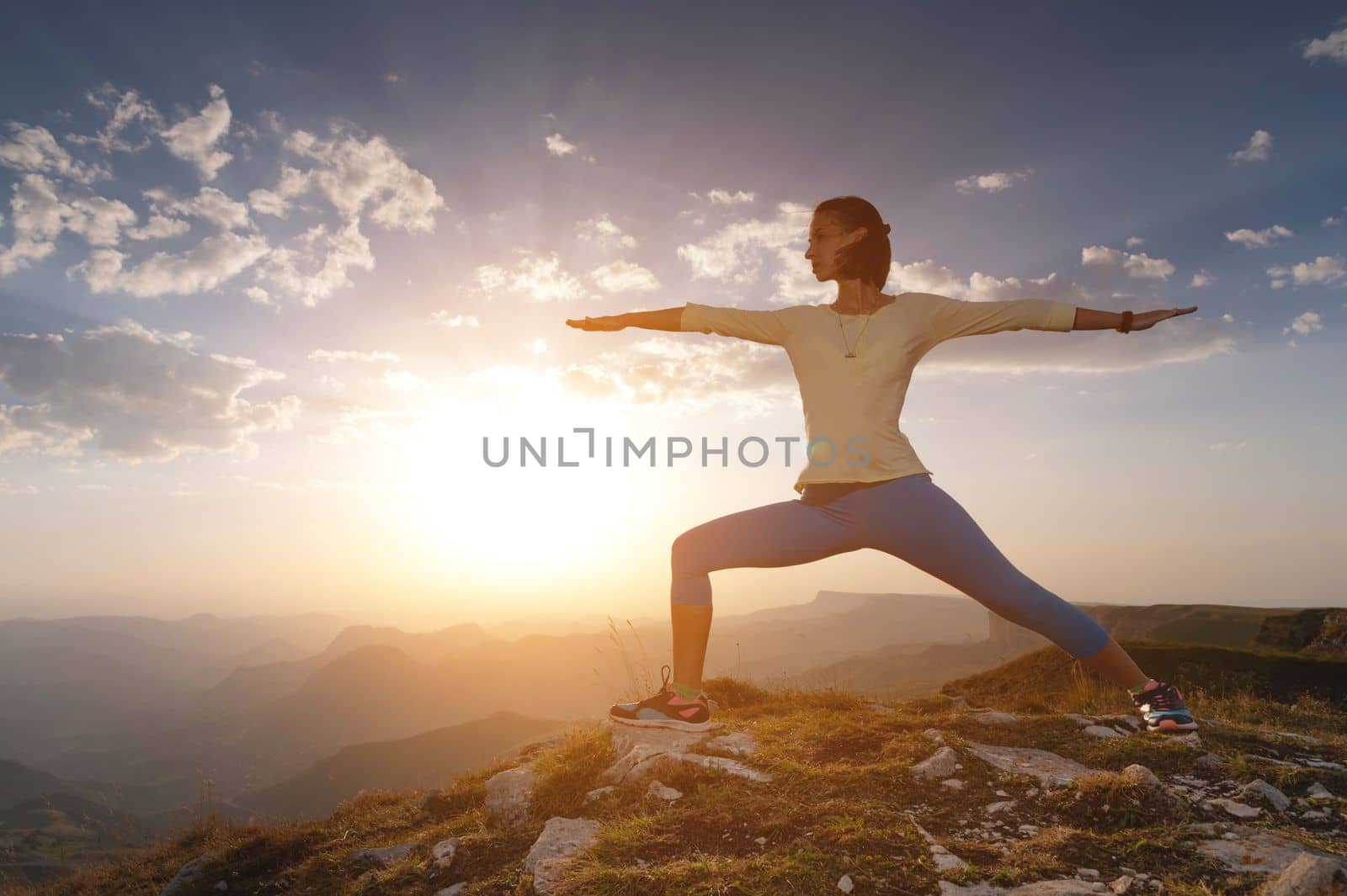 Young woman in warrior yoga pose standing on mountain rock under beautiful cloudy sky.