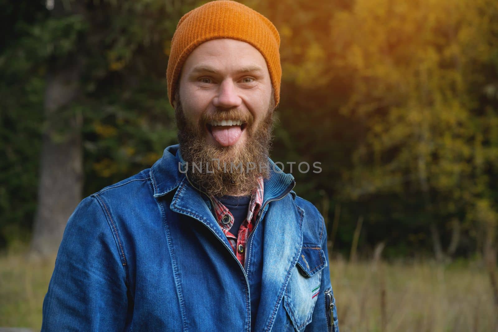 An attractive bearded man in a red hat and a denim jacket grimaces showing his tongue at the camera in the forest. funny portrait by yanik88