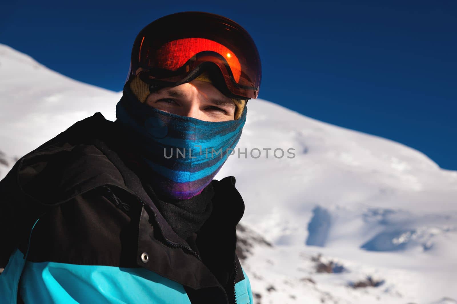 Portrait of a skier in the mountains without a ski mask with a scarf on his face, narrowing his eyes, looks at the camera against the backdrop of snow-capped mountains.