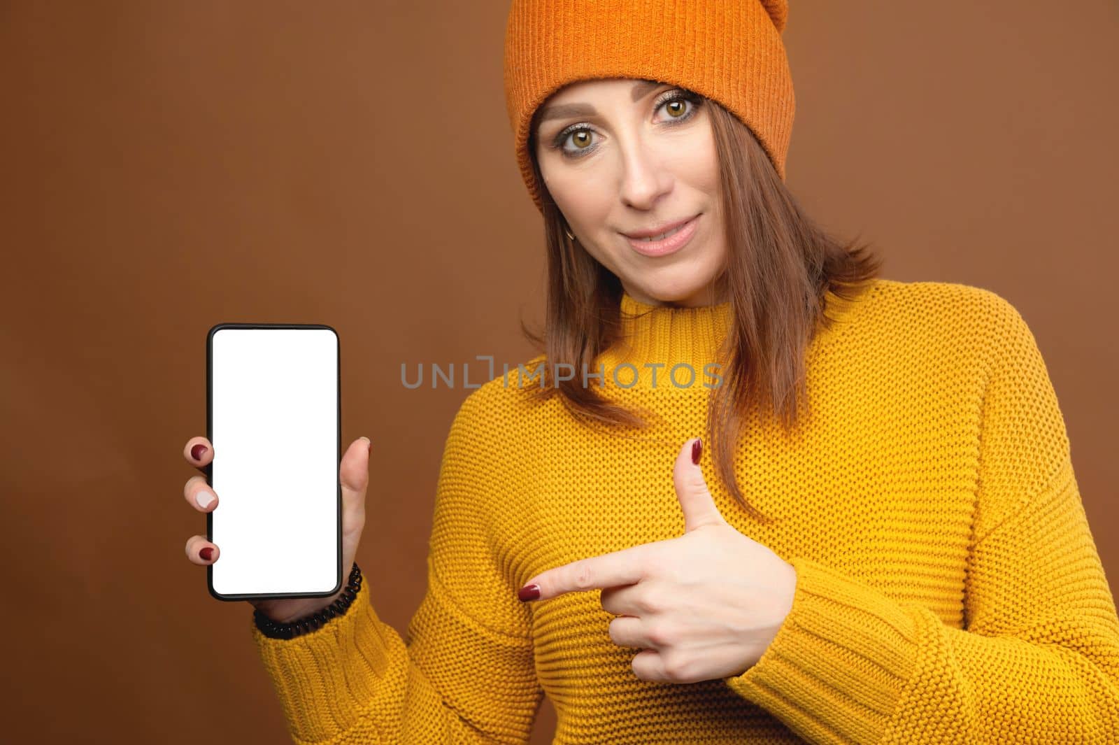 Middle-aged caucasian woman in a hat and sfiter holds a phone with a cut-out screen. points at it with a finger while looking at the camera.