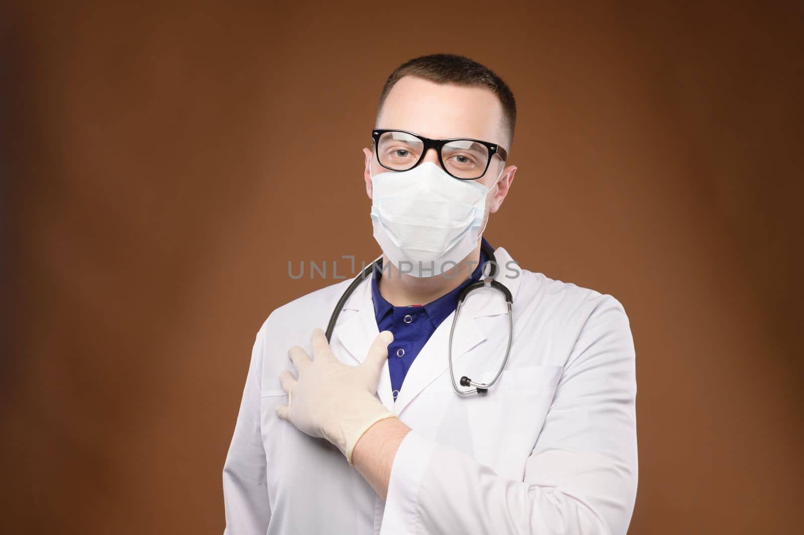 doctor in a medical mask, with a stethoscope around his neck and wearing gloves, stands in a white coat and holds his hand on his heart. Portrait of a doctor by yanik88