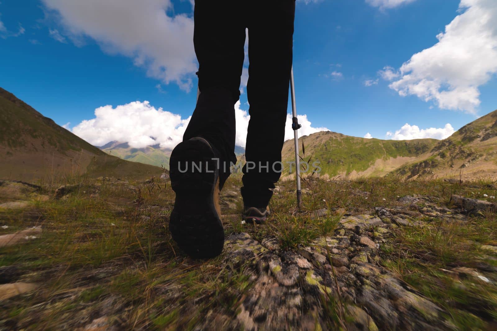 Legs of an athletic woman in black trousers and trekking boots with trekking poles walks high in the mountains.