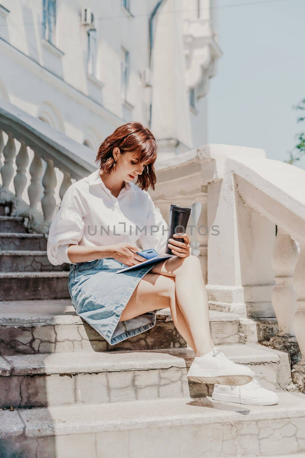Freelancer woman phone. A business woman in a white shirt and denim skirt sits on the steps near an ancient building in the city and looks into the phone by Matiunina
