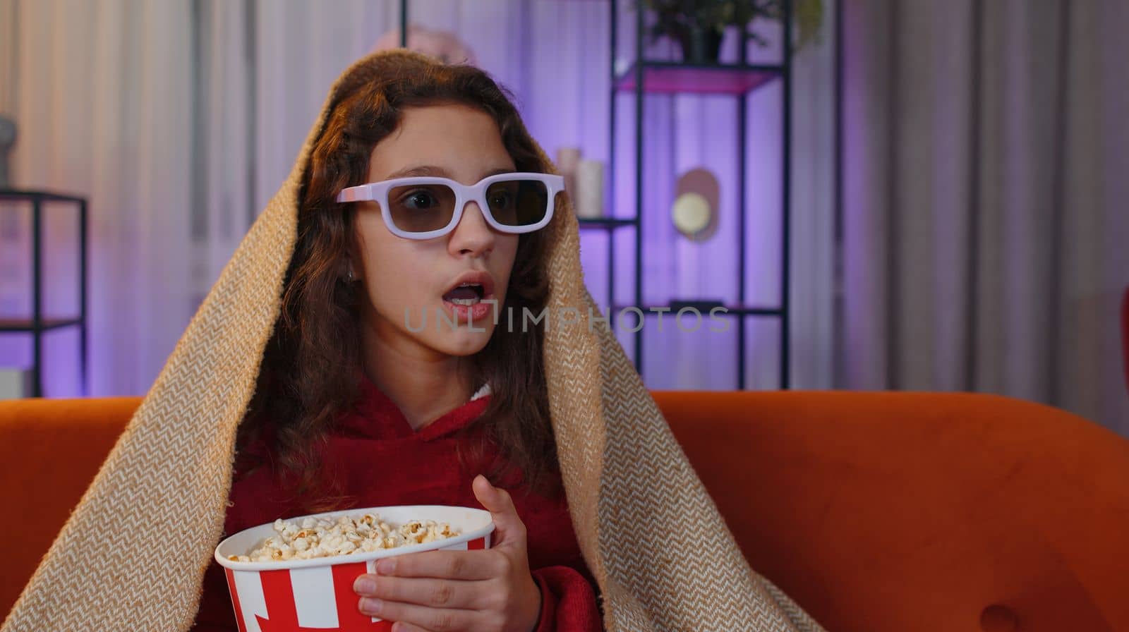 Excited preteen girl in 3D glasses eating popcorn and watching interesting tv serial, sport game, film, online social media movie content. Young child kid at home in evening living play room on couch