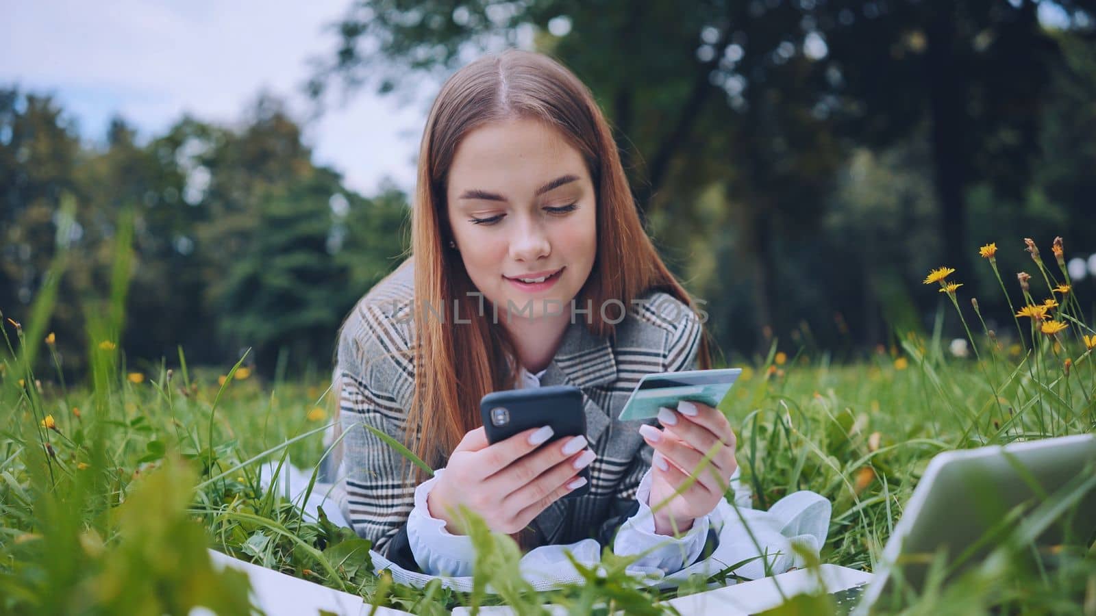 A young girl makes a purchase with her phone and bank card while lying on the grass in the park