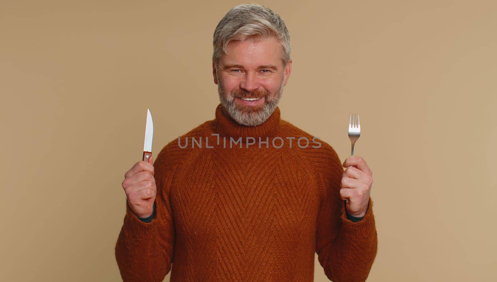 Ready to eat. Hungry middle-aged man with fork, knife waiting for serving dinner dishes with cutlery, will appreciate delicious restaurant meal lunch. Senior mature guy on beige studio background