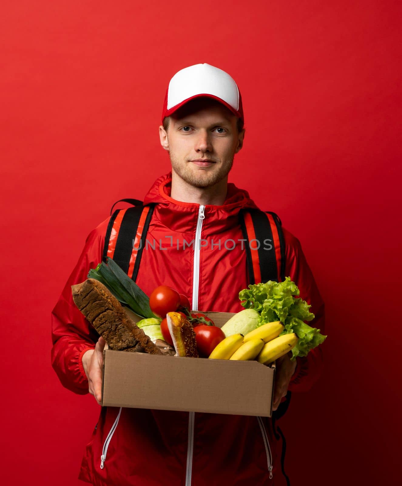 Close up portrait of delivery man in red uniform delivering food, groceries, vegetables, drinks in a paper box to a client at home. Online grocery shopping service concept