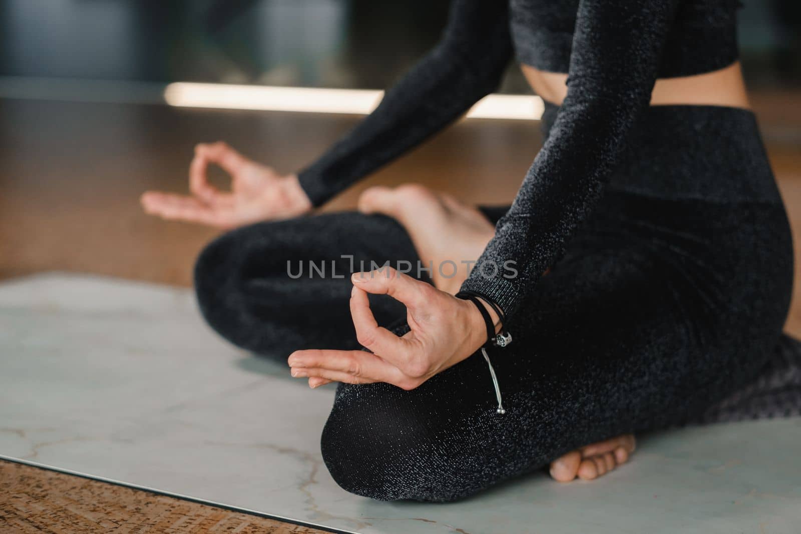 Close-up of the legs and arms of a woman in a black tracksuit sitting on a yoga mat in a lotus position in the gym.