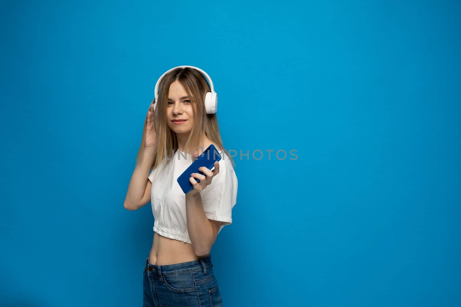Happy blonde woman girl wearing headphones listening to music from mobile phone and relaxing on isolated blue studio background