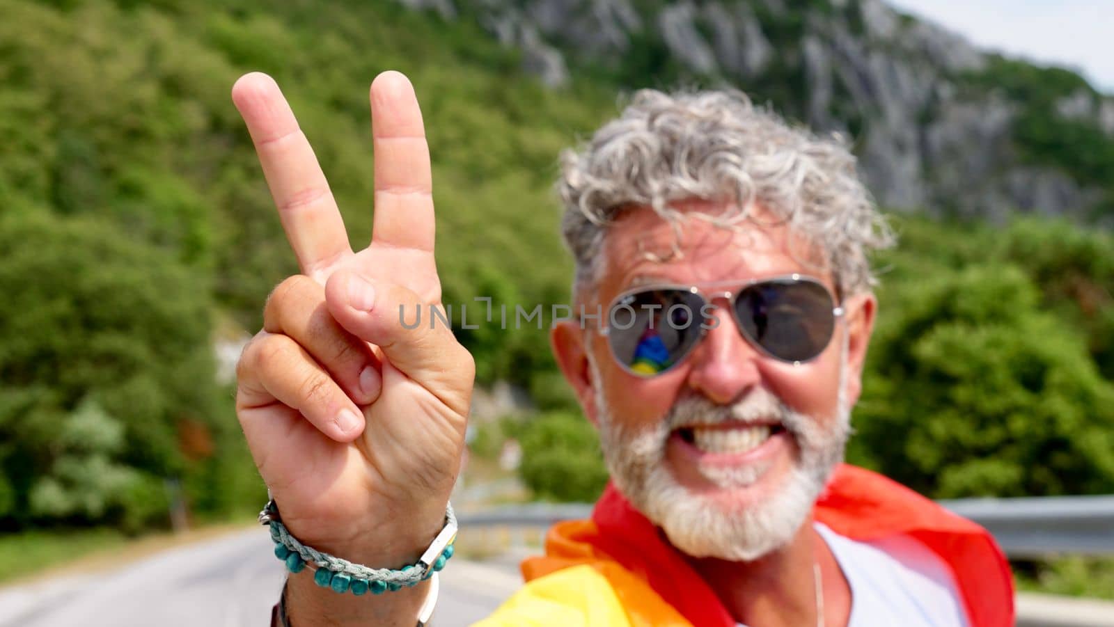 Portrait of a gray-haired senior elderly Caucasian man bisexuality with a beard and sunglasses with a rainbow LGBTQIA peace flag in mountains. Celebrates Pride Month, Rainbow Flag Day, gay parade