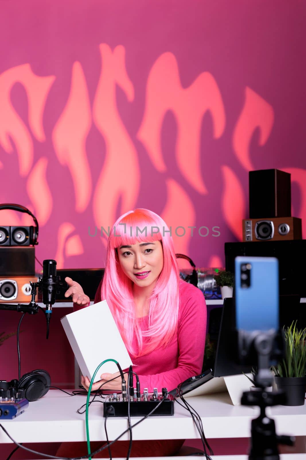 Content creator with pink hair holding white box presenting product in front of phone camera, filming review for vlogging channel. Asian influencer recording vlog using professional vlogging equipment