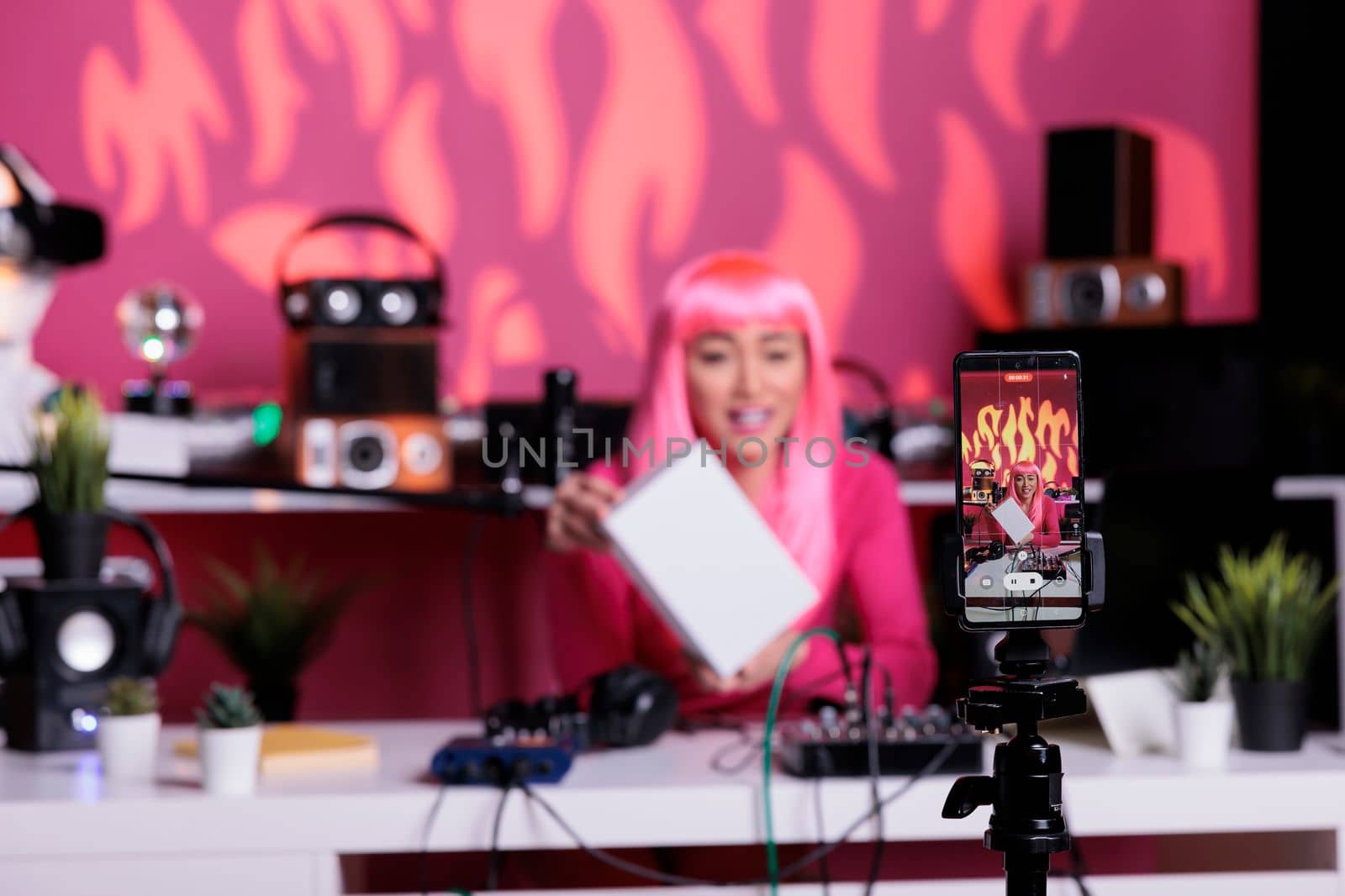 Asian woman with pink hair holding white box advertising product while recording vlog using smartphone camera in broadcast studio. Influencer filming content using professional equipment