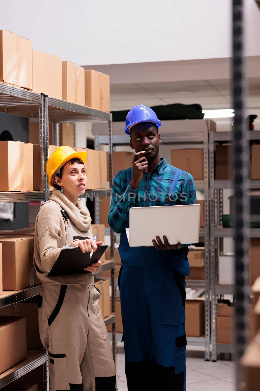 Warehouse workers checking factory goods supply and monitoring dispatching operations on laptop. Caucasian and african american storehouse colleagues standing near cardboard boxes shelf