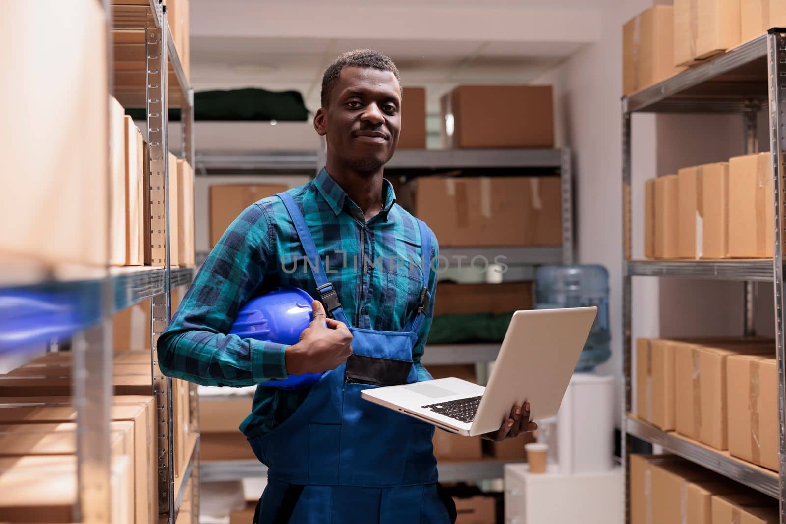 Warehouse employee doing inventory management using laptop, checking parcels before transportation and looking at camera. Smiling african american storehouse worker portrait