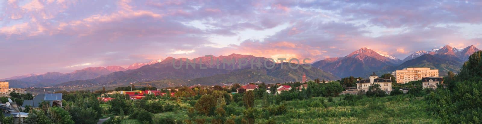 Panorama of the city of Almaty immersed in summer greenery against the backdrop of mountains in sunset lighting. Almaty, Kazakhstan - June 18, 2022