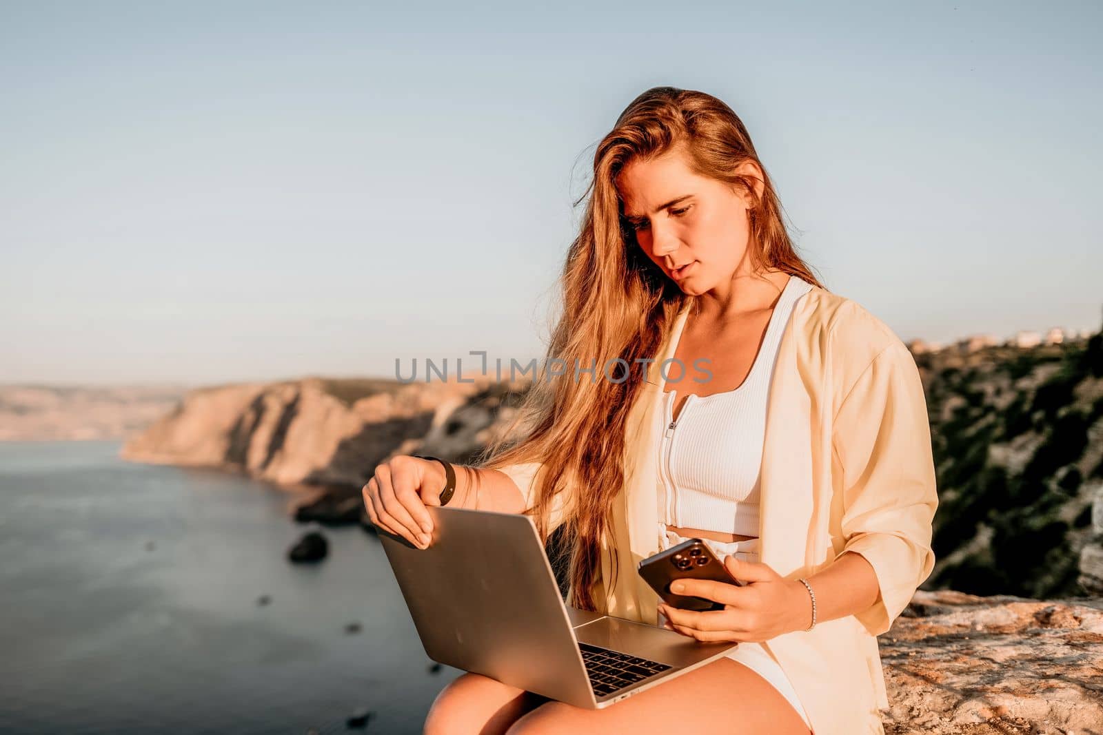 Successful business woman in yellow hat working on laptop by the sea. Pretty lady typing on computer at summer day outdoors. Freelance, travel and holidays concept.