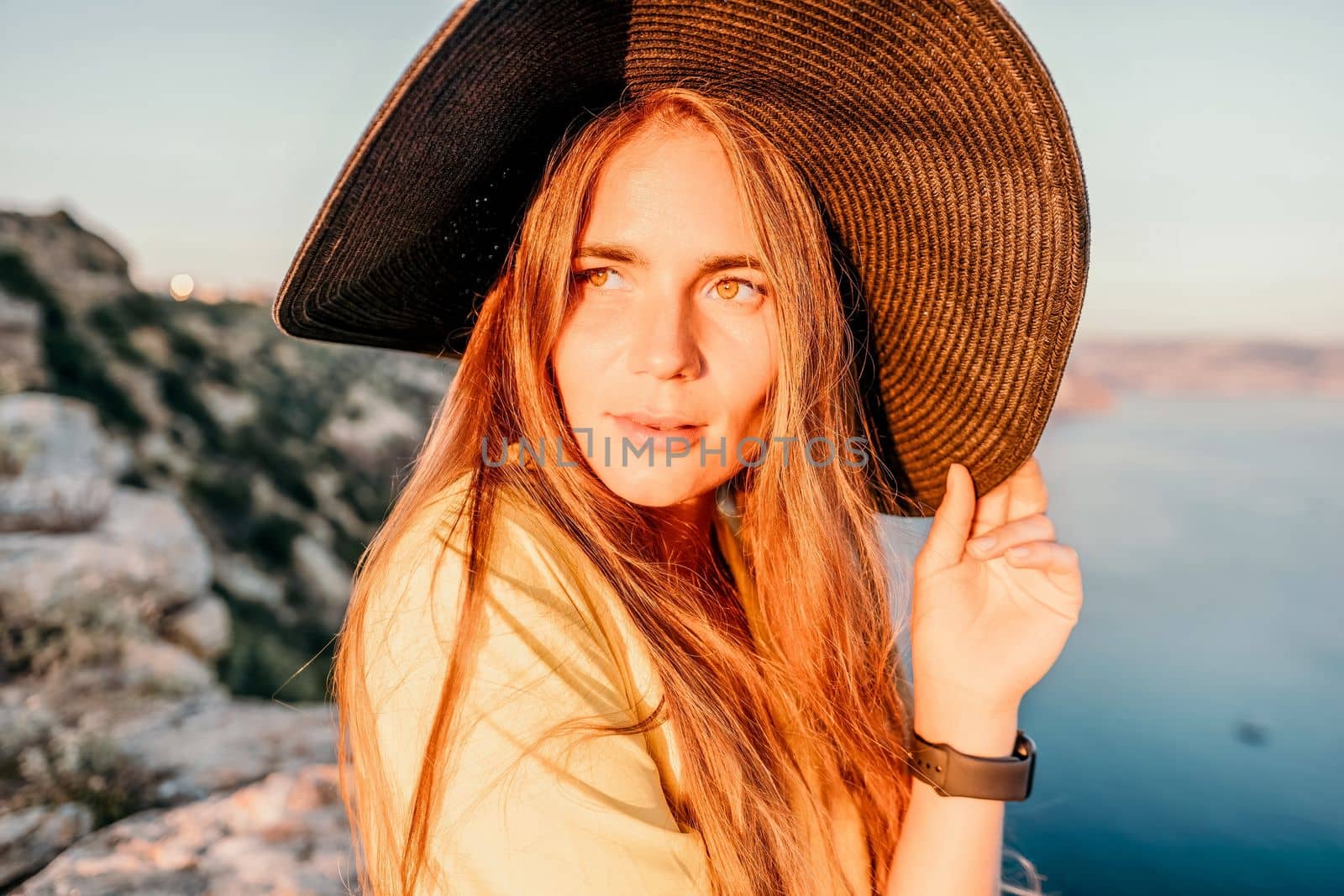 Portrait of happy young woman wearing summer black hat with large brim at beach on sunset. Closeup face of attractive girl with black straw hat. Happy young woman smiling and looking at camera at sea
