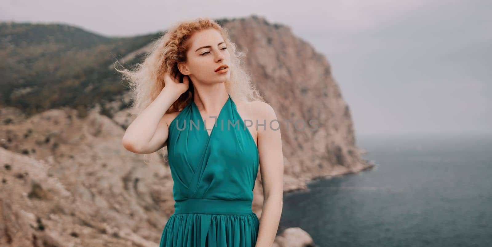 Side view a Young beautiful sensual woman in a mint long dress posing on a volcanic rock high above the sea during sunset. Girl on the nature on overcast sky background. Fashion photo