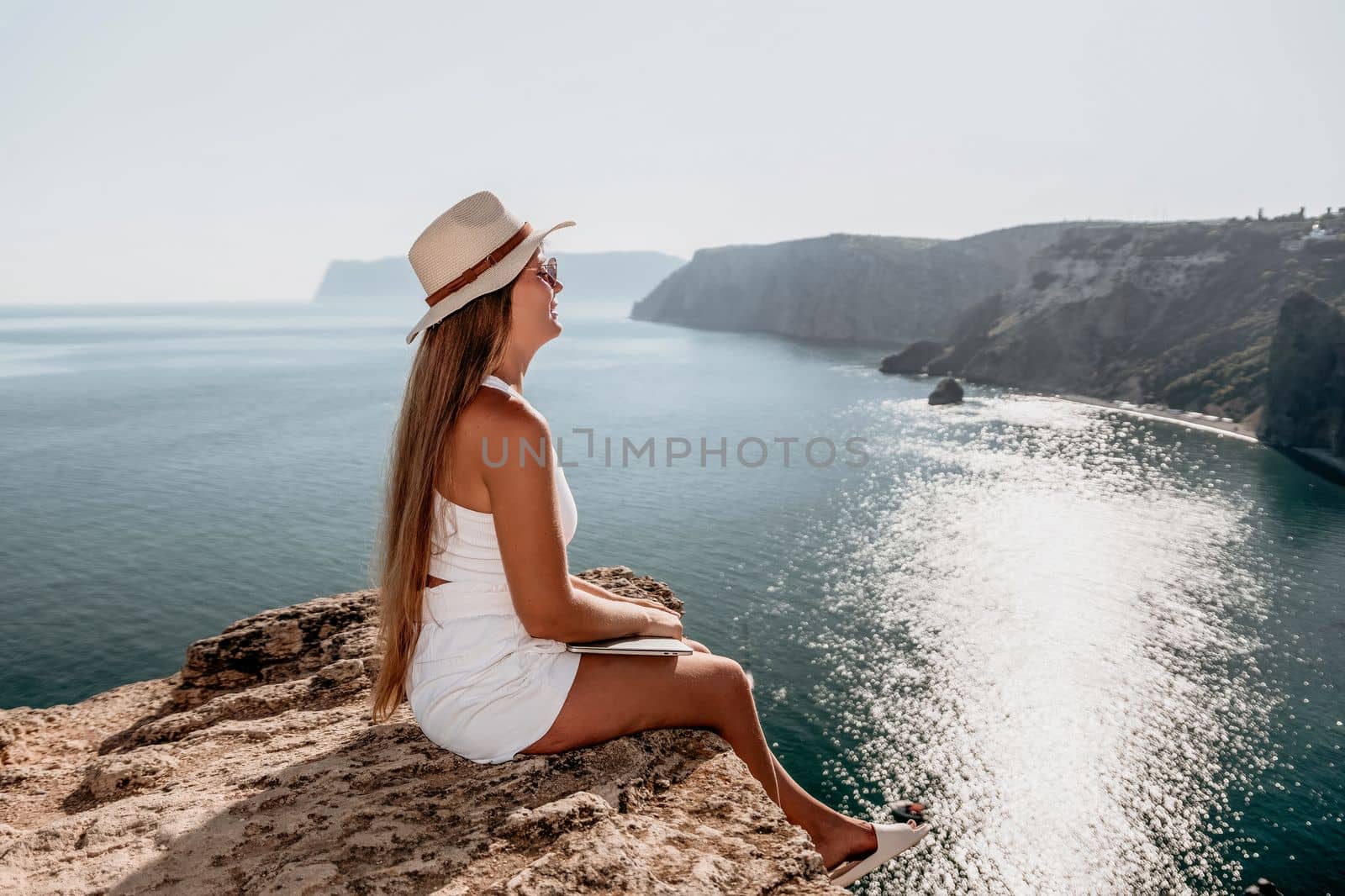 Successful business woman in yellow hat working on laptop by the sea. Pretty lady typing on computer at summer day outdoors. Freelance, travel and holidays concept.