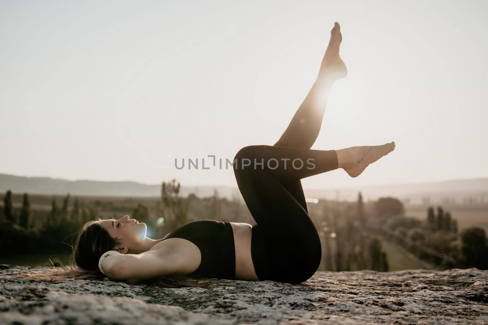 Well looking middle aged woman with long hair, fitness instructor in leggings and tops doing stretching and pilates on the rock near forest. Female fitness yoga routine concept. Healthy lifestyle.
