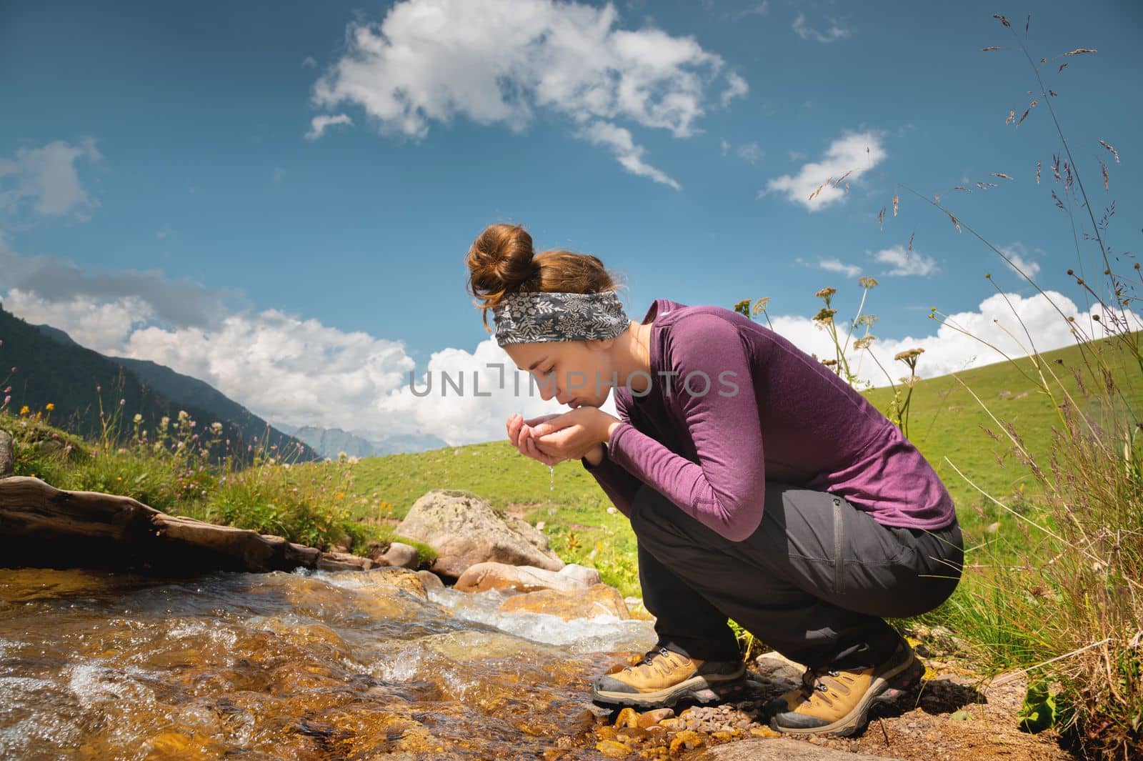 A woman tourist takes water in the river after a walk. Happy girl smiling while enjoying summer holidays outdoors in the mountains.