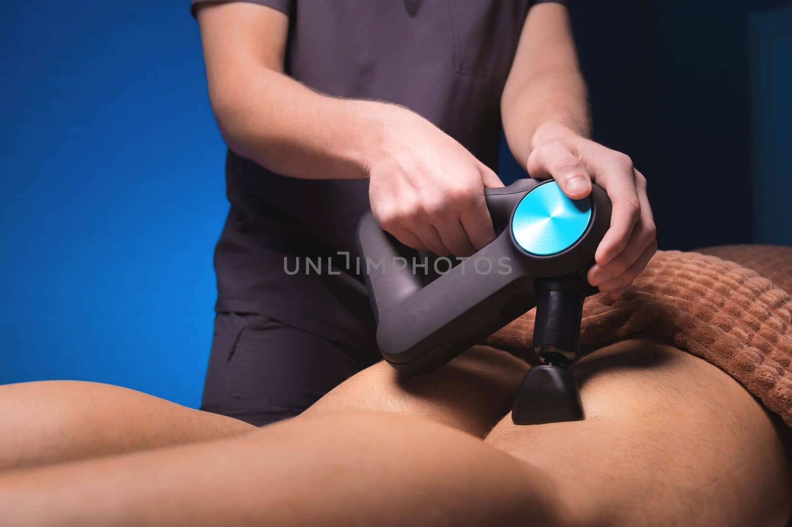Close-up of unrecognizable professional male masseur massaging leg calf muscles using massage gun percussion tool of muscular athlete man, on spa treatment lying on back in massage table.