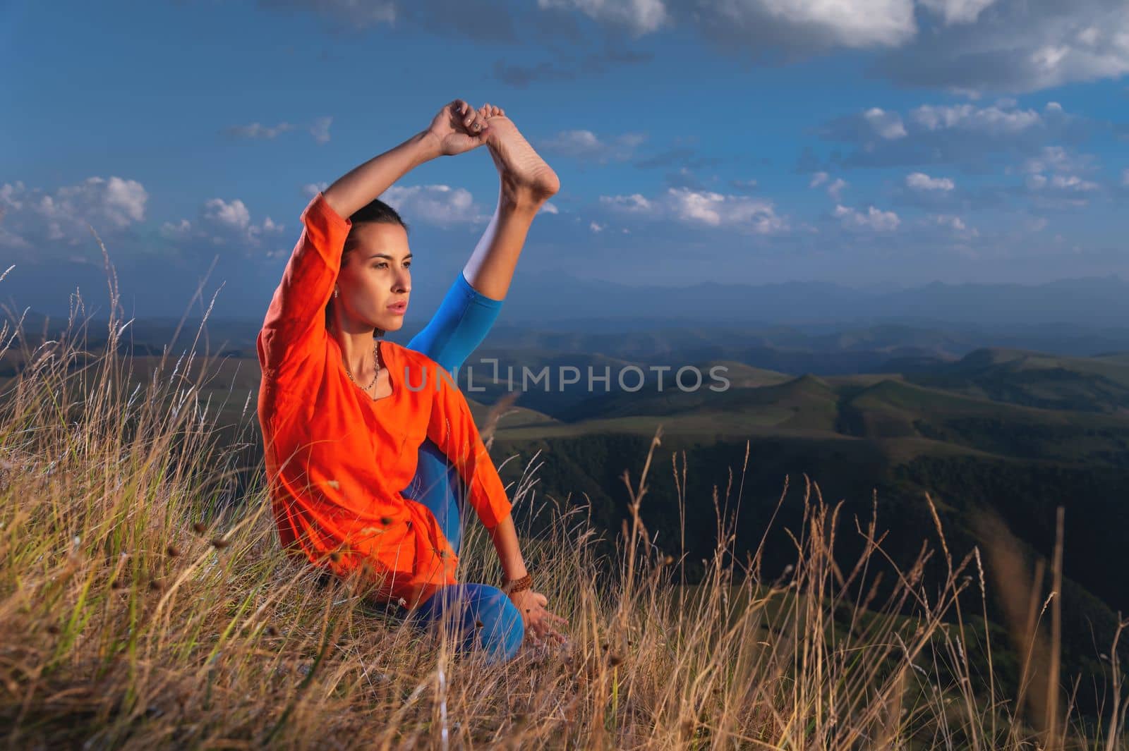 young woman practicing yoga poses raising one leg to her head while sitting on the rocks in the sunset light in the mountains.