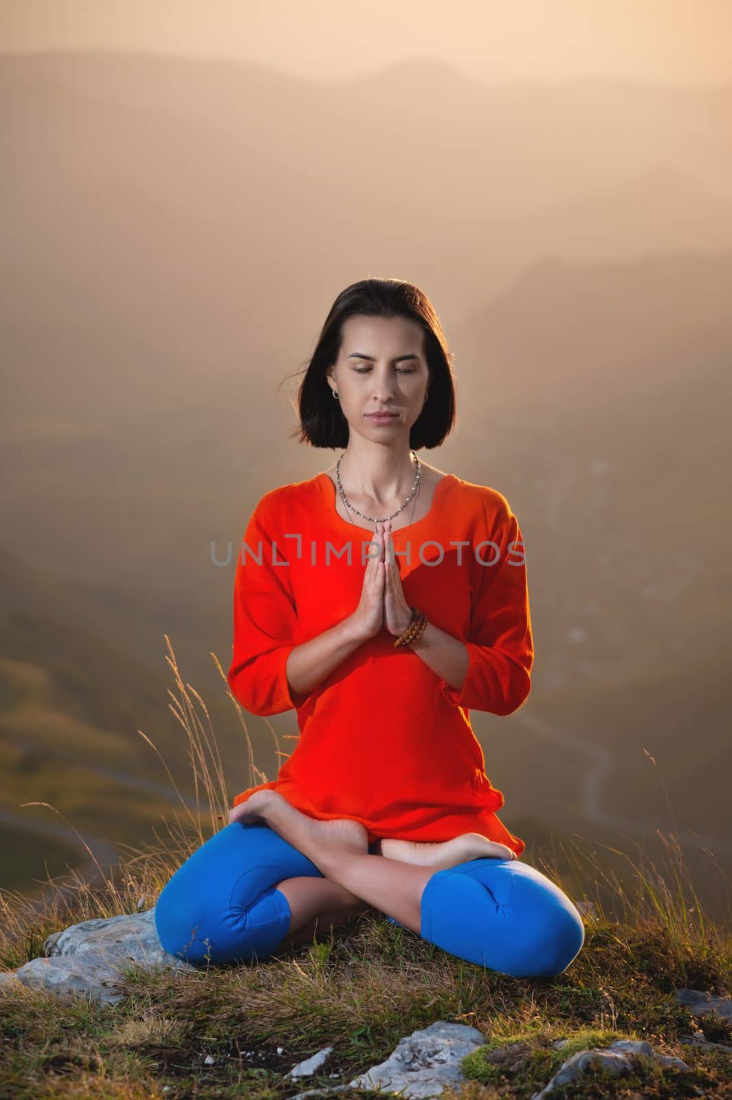 young caucasian woman meditates in the lotus position in the grass in the mountains near the cliff in the setting sun.