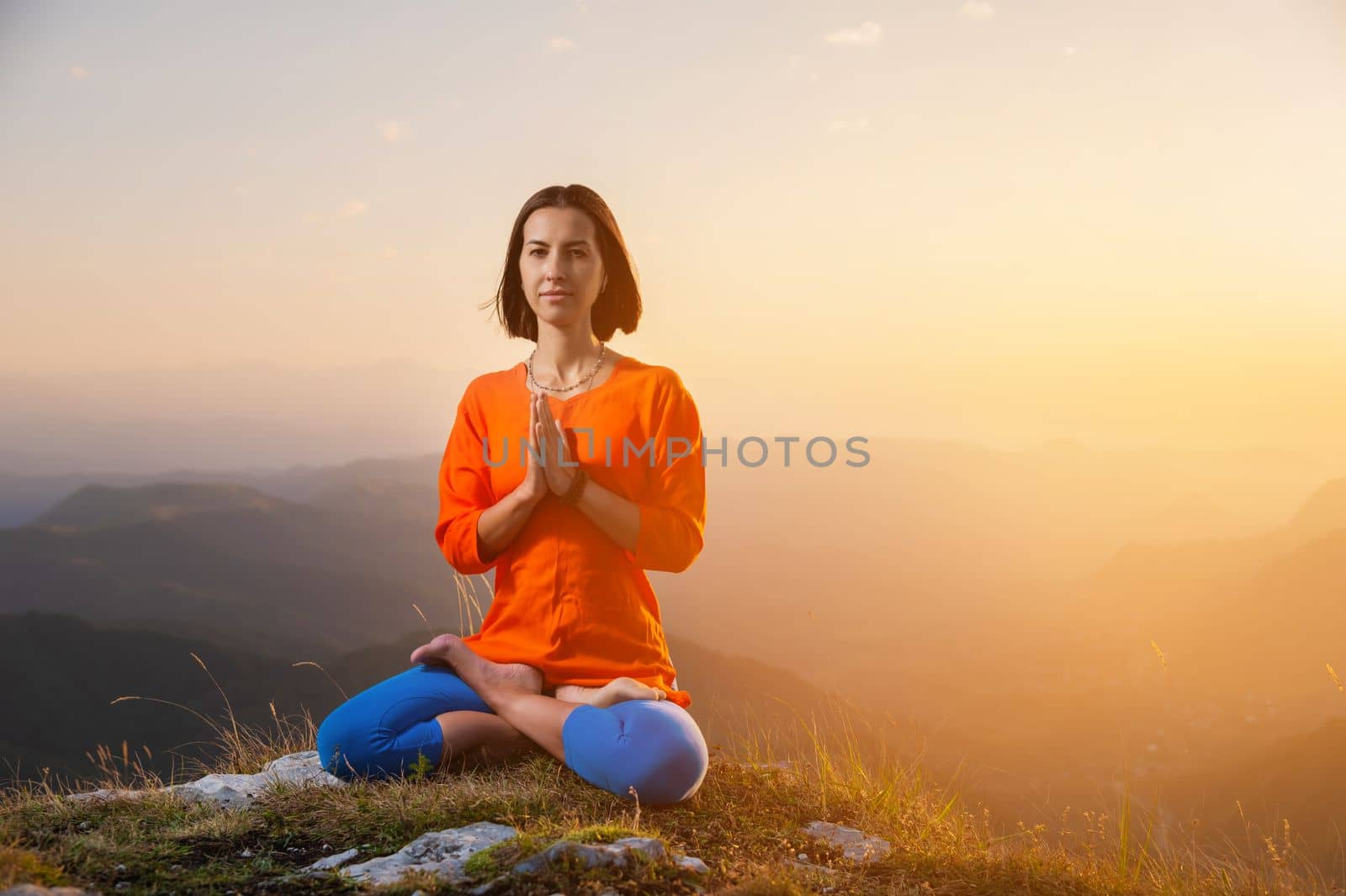 a yogi woman sits in a lotus position with crossed legs and holds her hands to her chest against the backdrop of a sunset in the mountains, there is a place for advertising on the side.