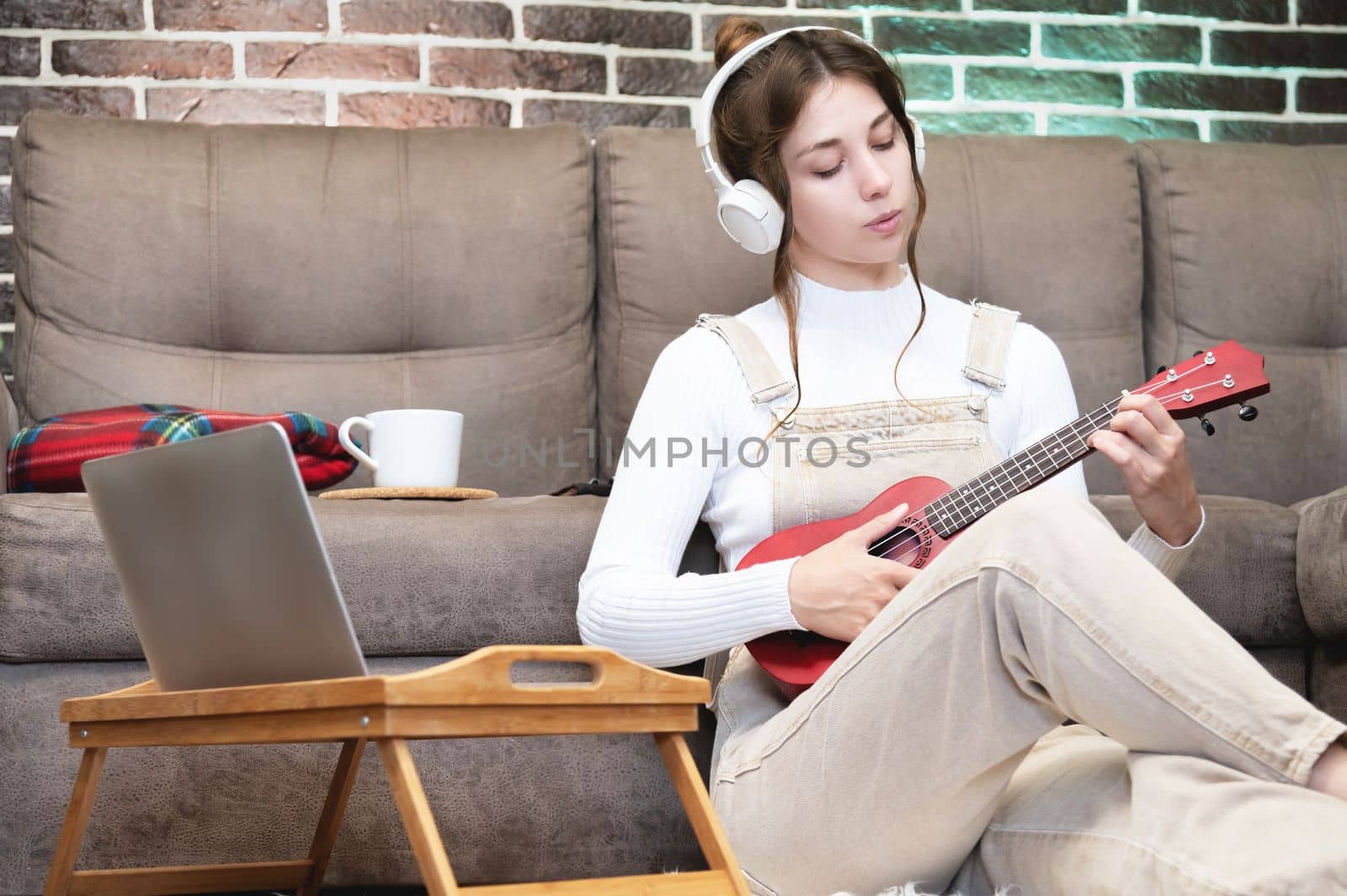 Beautiful young hipster girl in casual clothes learning to play the ukulele guitar while sitting on the floor in the living room at home, hobby leisure time concept.