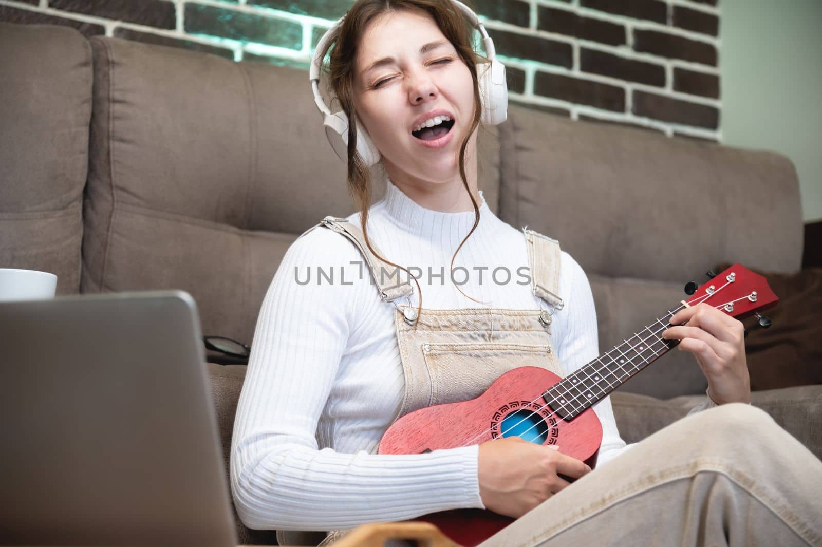 young woman student learning vocals and playing the ukulele at home sitting on the floor in the living room in front of a laptop.