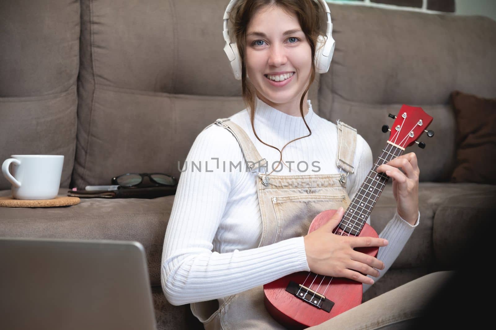 Beautiful young hipster girl in casual clothes learning to play the ukulele guitar while sitting on the floor in the living room at home, hobby leisure time concept. look at camera