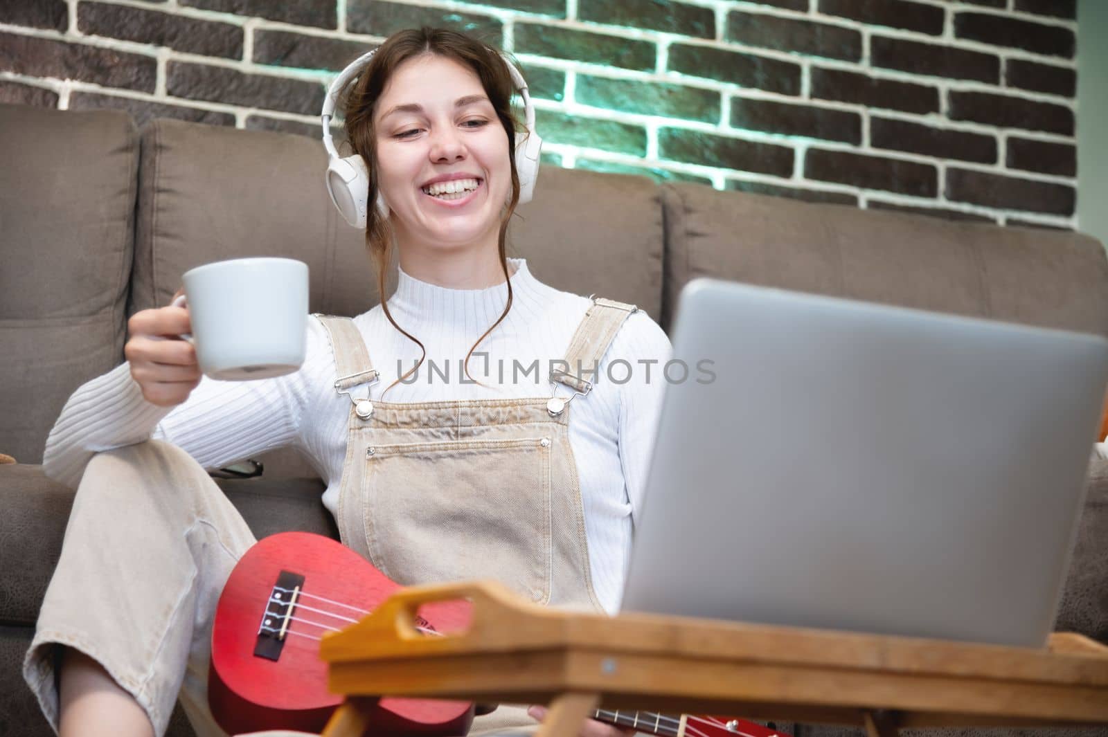 happy young woman sitting with a cup of coffee and ukulele, wearing headphones on the floor in the living room in her house, looking into the laptop monitor, distance learning.