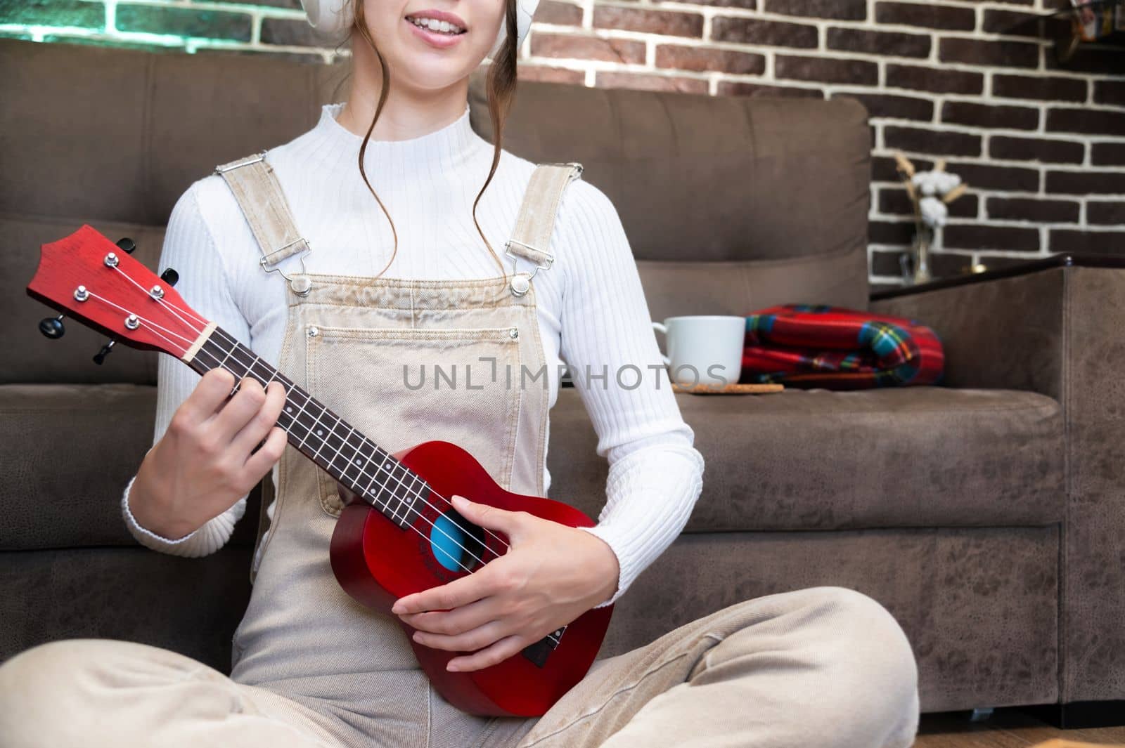 close-up of female hands playing the red ukulele sitting at home against the background of the sofa.