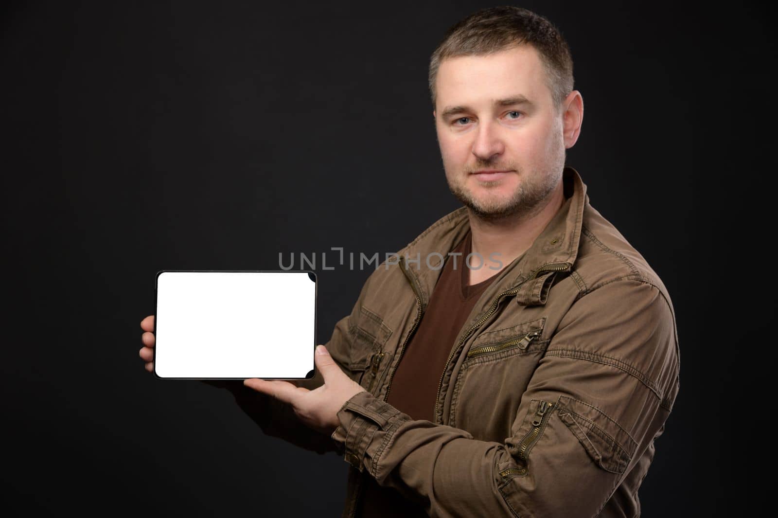 Smiling unshaven business man in a leather jacket on a black wall background. Achieving career wealth business concept. Holding tablet pc computer with a cut out screen.