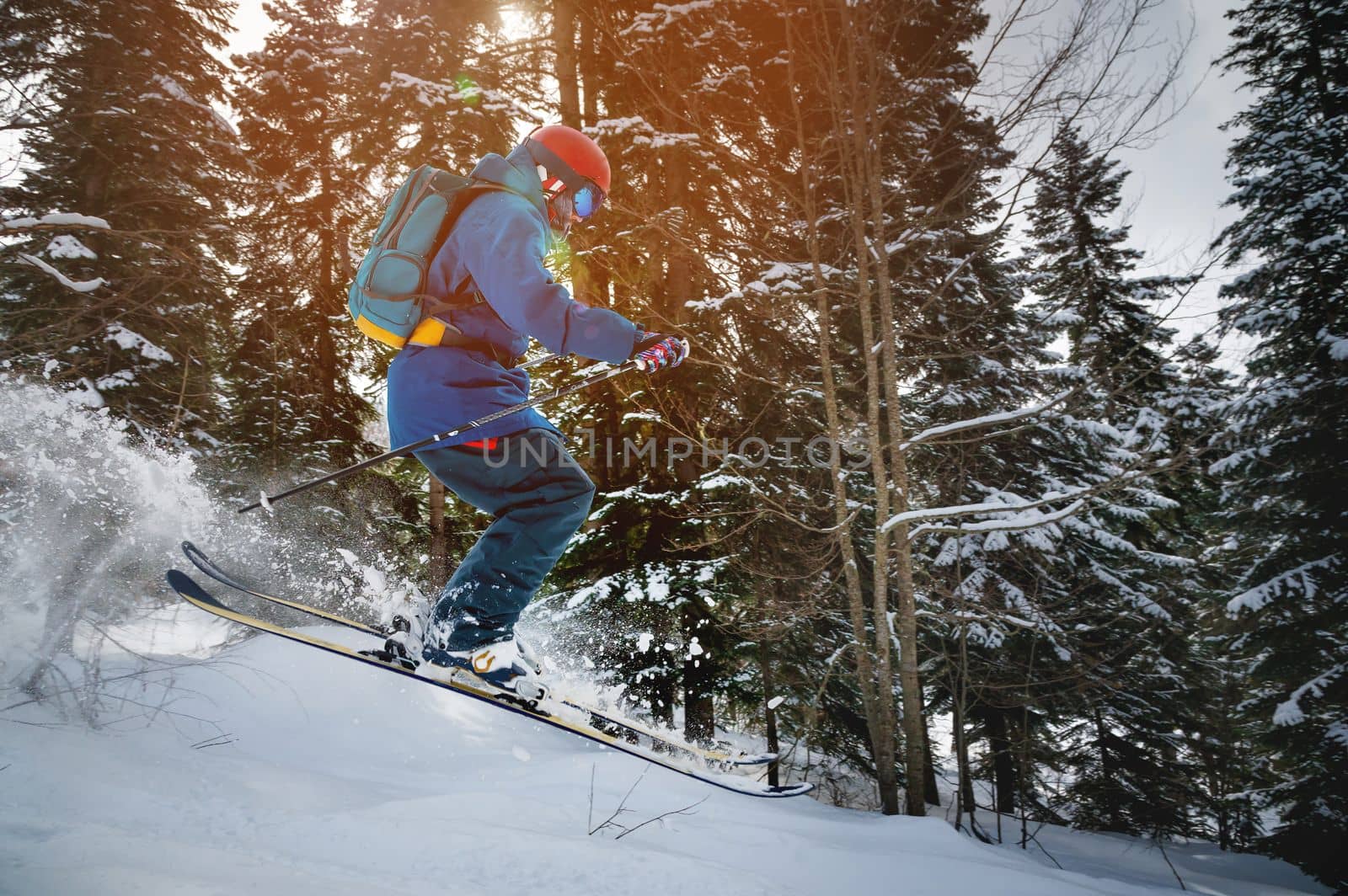 a skier makes a ski jump with a plume of snow behind him, in the mountains against the backdrop of a dense forest. male athlete in bright clothes makes a downhill race.