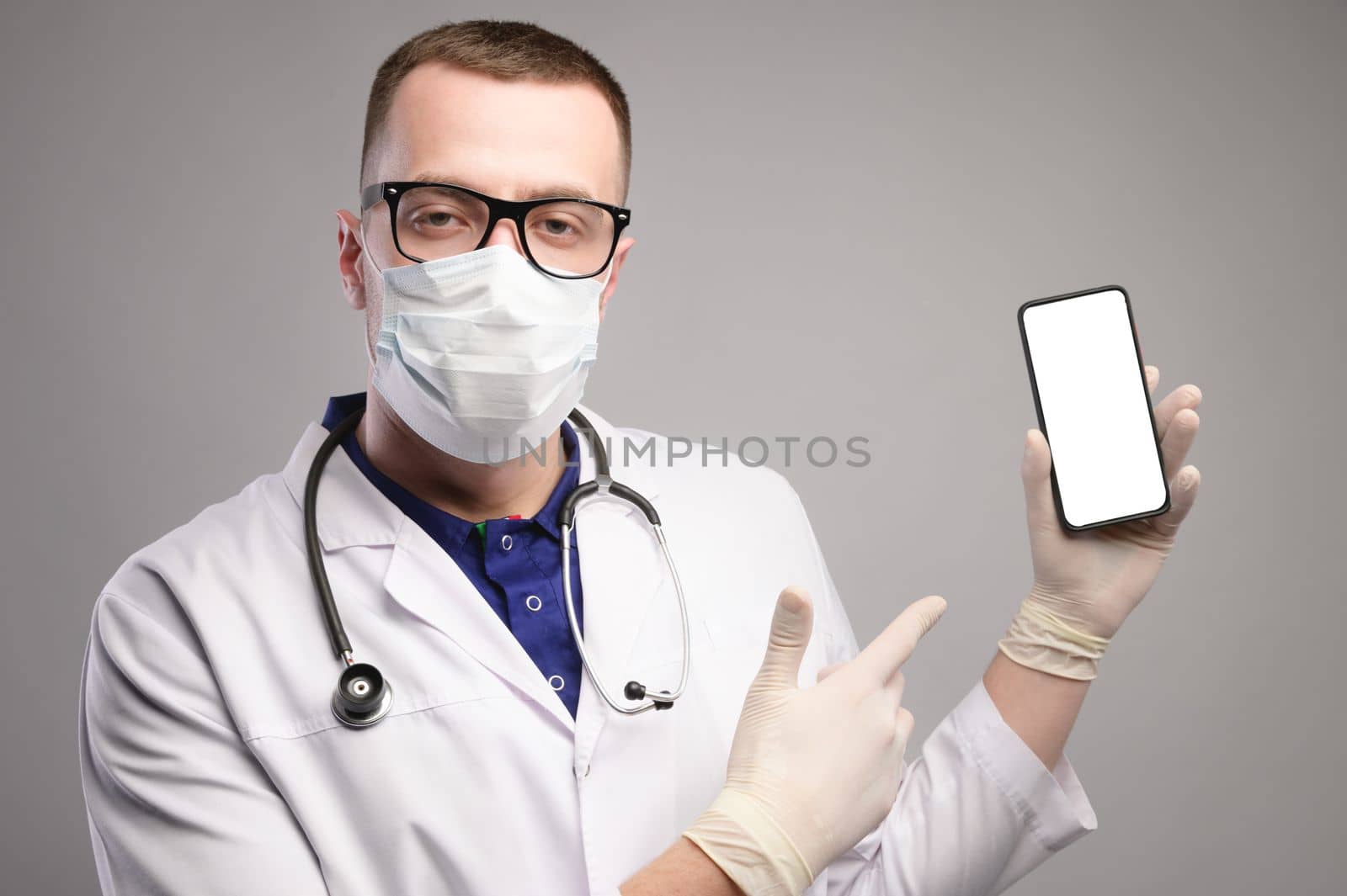 a young doctor in glasses and a mask stands pointing at the smartphone with his finger, looking at the camera, a portrait in a bathrobe.