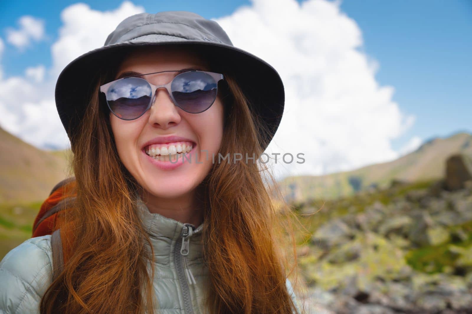 Smiling young girl against a blue cloudy sky and mountains. Portrait of a happy woman tourist in sunglasses and panama.