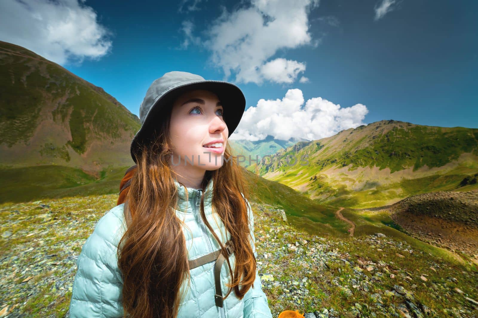 Smiling, positive girl in full face against a blue sky with white clouds in the mountains, a happy portrait of a tourist with a backpack.