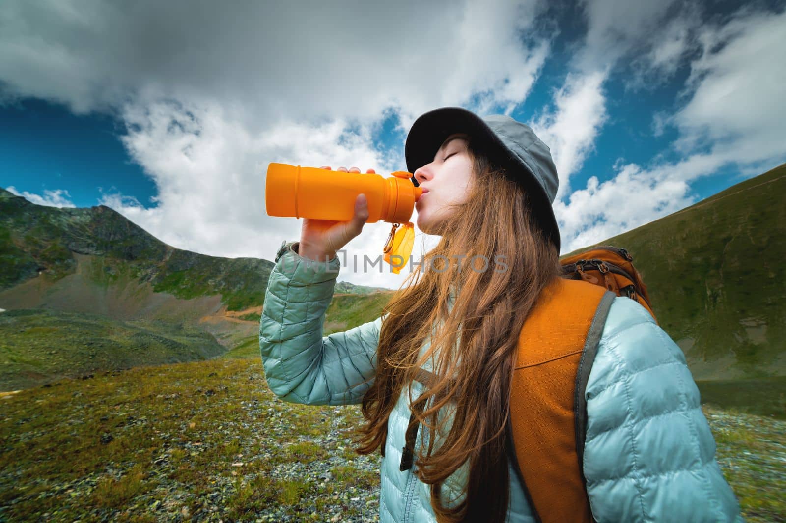 Profile photo of a young beautiful woman with long blond hair, dressed in a silver jacket, drinking from a yellow camping bottle, in the background a magnificent landscape of mountains and clouds by yanik88
