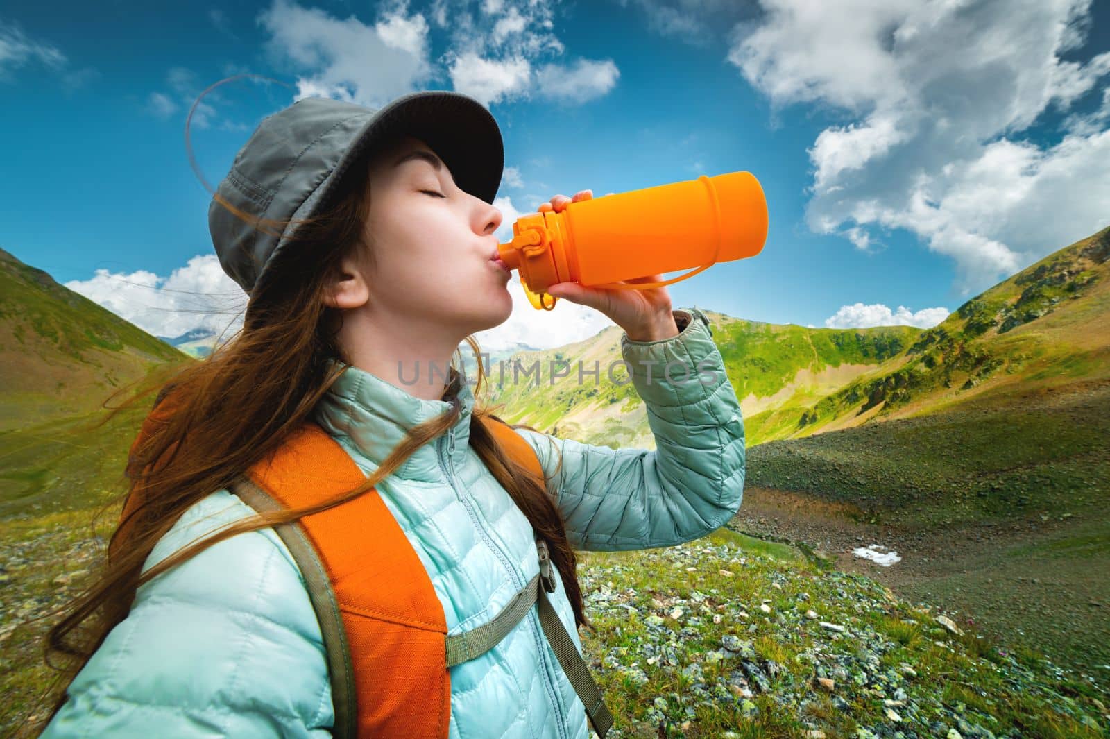 Young sports girl stands against the backdrop of a stunning mountain view and drinks clean water from a rubber bottle, trekking with a backpack.