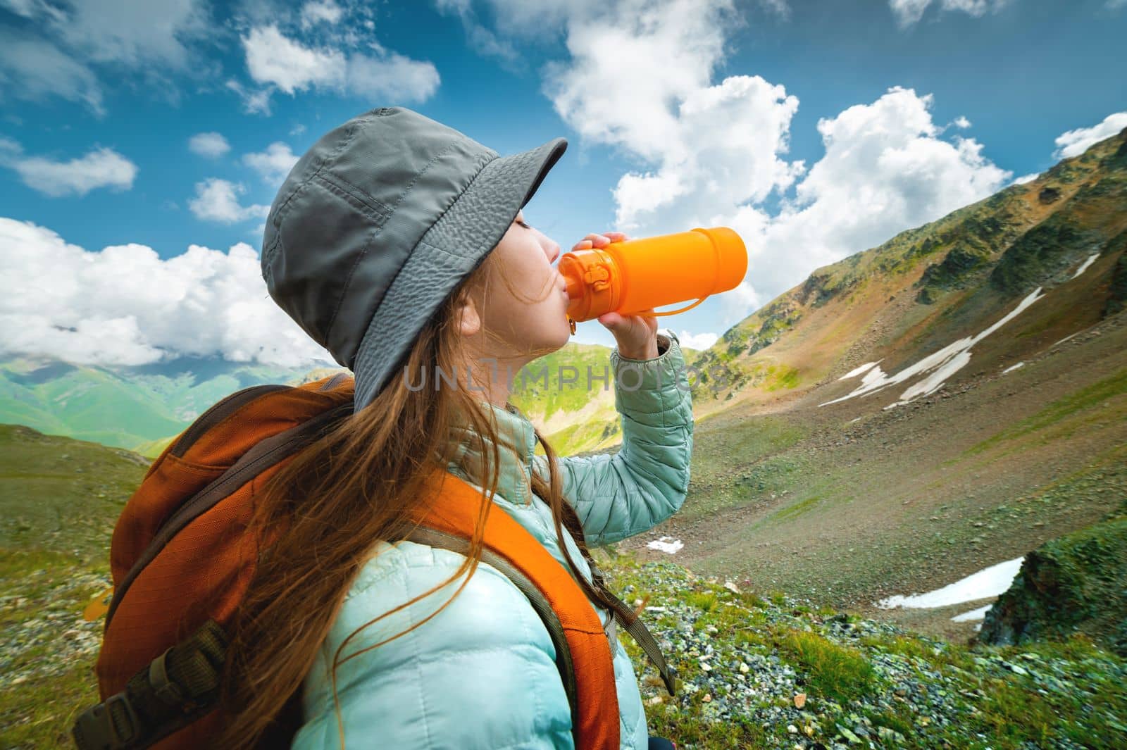 Profile photo of a young beautiful woman with long blond hair, dressed in a silver jacket, drinking from a yellow camping bottle, in the background a magnificent landscape of mountains and clouds.