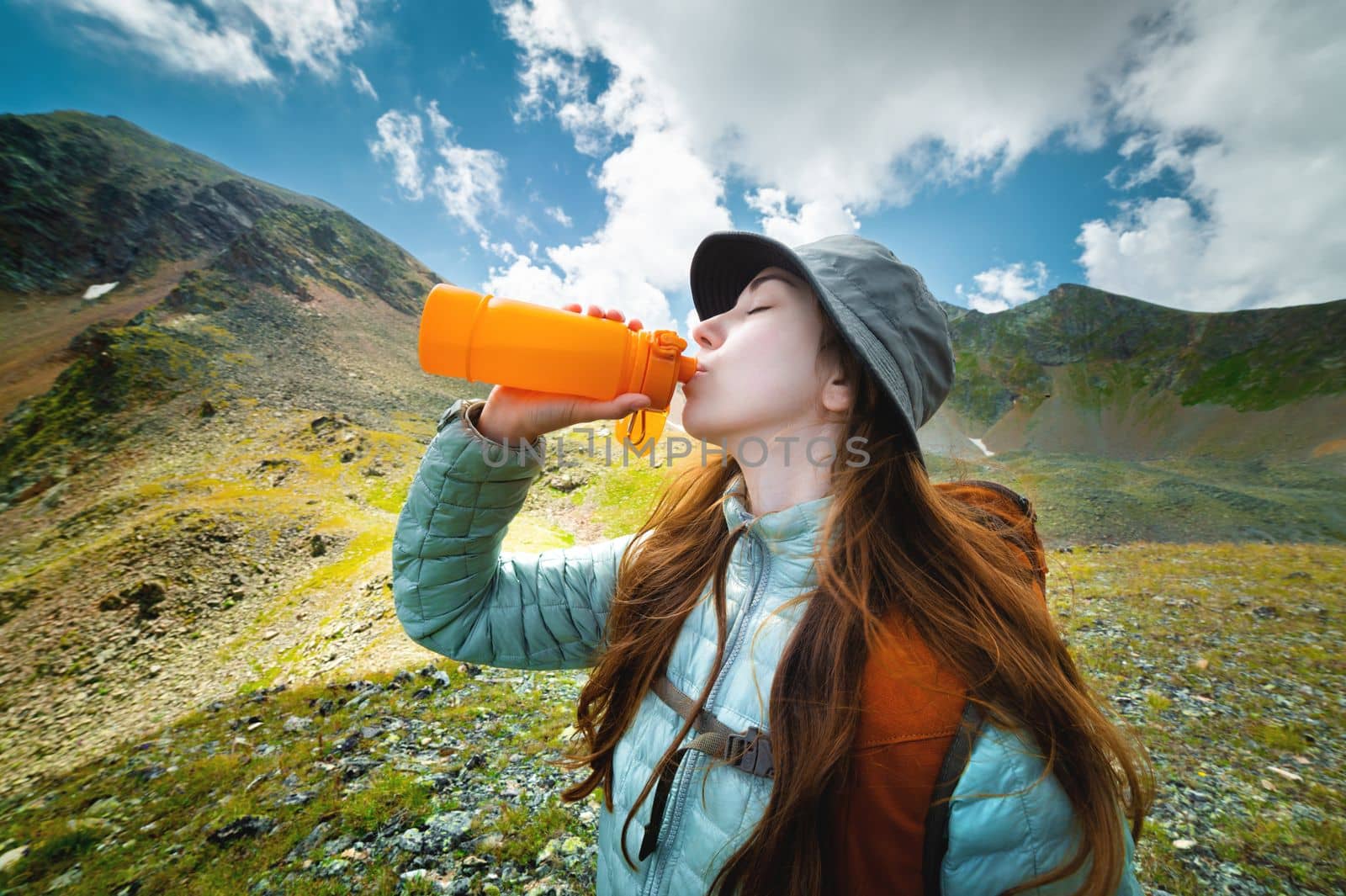 Profile photo of a young beautiful woman with long hair, wearing a jacket and a panama hat with a backpack, drinking from a yellow bottle, against the backdrop of a magnificent mountain landscape.