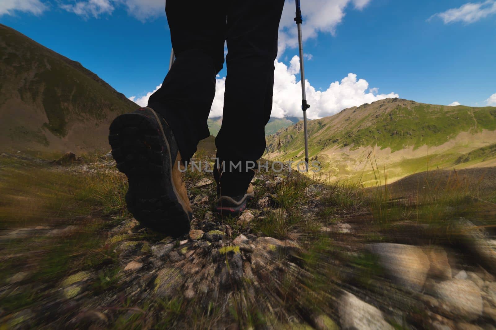 close-up, legs in trekking shoes with sticks walk along the stones to the lake against the backdrop of mountains, view from back in motion.