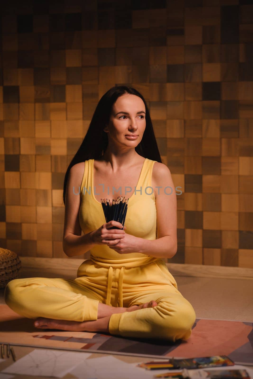 A girl in yellow clothes sitting on the floor holding pencils for drawing.