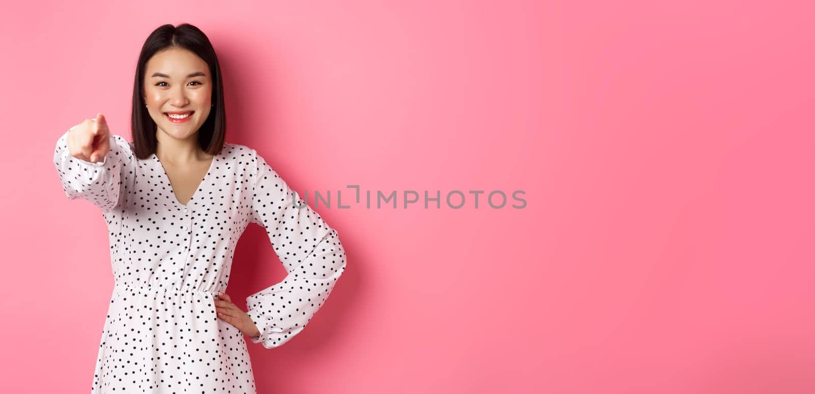 Confident asian woman in dress pointing at you, smiling and staring at camera, standing against pink background by Benzoix